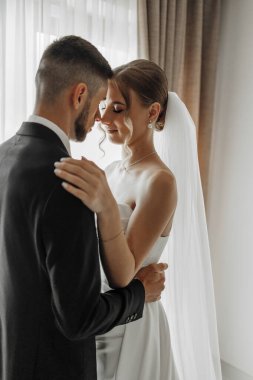 Bir gelin ve damat düğün fotoğrafında birbirlerine sarılıyorlar. Gelin beyaz bir elbise giyiyor ve damat bir takım elbise giyiyor. Sahne mutlu ve romantik, çift aşklarını kutluyor.