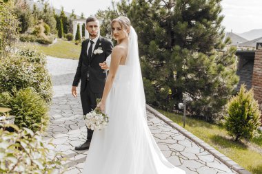 Bir gelin ve damat beyaz bir gelinlik ve beyaz bir buketle bir evin önünde duruyorlar.