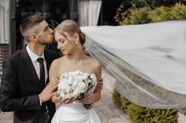 Bir gelin ve damat birbirlerinin yanaklarını öpüyor. Gelin elinde bir buket beyaz çiçek tutuyor.