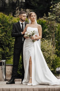 Bir gelin ve damat bir çalılığın önünde fotoğraf için poz veriyorlar. Gelin elinde bir buket çiçek tutuyor.