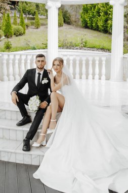 Bir gelin ve damat beyaz mermer bir merdivende fotoğraf için poz veriyorlar. Gelin beyaz bir elbise giyiyor ve damat siyah bir takım elbise giyiyor. Sahne mutlu ve romantik.