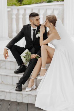 Gelin ve damat merdivenlerde oturuyor, gelin beyaz bir elbise giyiyor ve damat siyah bir takım giyiyor.