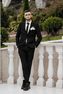 Takım elbiseli ve kravatlı bir adam balkonda duruyor. Yakasına beyaz bir çiçek takmış.