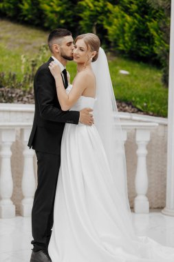 Bir gelin ve damat beyaz bir gelinlik ve takım elbise içinde birbirlerine sarılıyorlar. Gelin peçe takıyor ve damat yüzük takıyor. Sahne mutlu ve romantik.