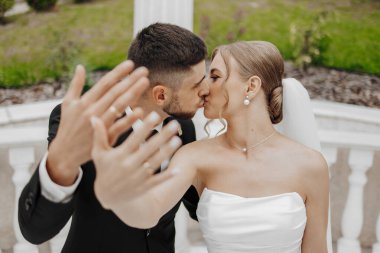 Bir gelin ve damat düğün fotoğrafında birbirlerinin ellerini öperler. Gelin beyaz bir elbise giyiyor ve damat da takım elbise giyiyor.