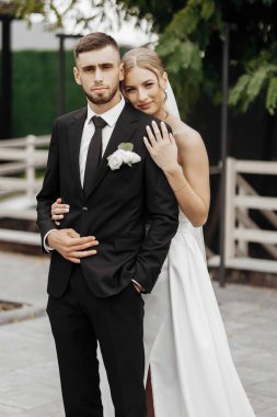 Bir gelin ve damat çitin önünde fotoğraf için poz veriyorlar. Gelin beyaz bir elbise giyiyor ve damat siyah bir takım elbise giyiyor. Sahne mutlu ve kutlama dolu.