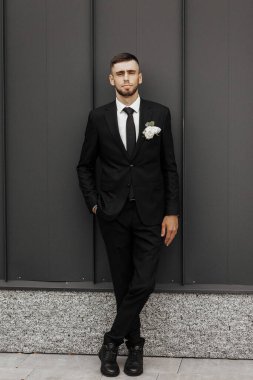 Siyah takım elbiseli ve beyaz kravatlı bir adam duvarın önünde duruyor. Elinde bir çiçek tutuyor.