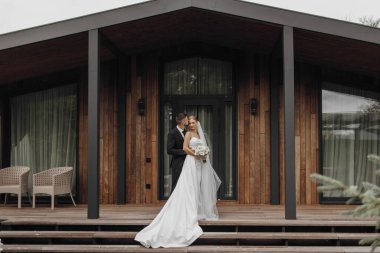 Gelin ve damat ahşap bir güvertede el ele tutuşup gülümsüyor. Gelin beyaz bir elbise giyiyor ve damat bir takım elbise giyiyor. Etrafları sandalyeler ve saksılarla çevrili.