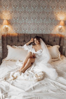 Beyaz elbiseli bir kadın bir yatakta oturuyor. Duvak ve alyans takıyor. Oda çiçeklerle süslenmiş ve sıcak, davetkar bir atmosferi var.