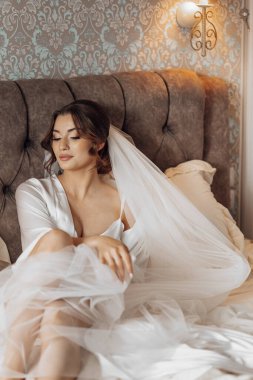 Gelinlik giymiş bir kadın kafasında peçeyle yatakta oturuyor. Zarafet ve romantizm kavramı, çünkü kadın gelinlik giyiyor ve büyük ihtimalle özel bir olaya hazırlanıyor.