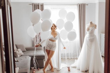 Bir kadın bir sürü beyaz balon tutuyor ve şifoniyerin önünde duruyor. Odanın rahat ve eğlenceli bir atmosferi var.