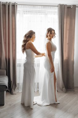 İki kadın düğün için hazırlanıyor. Biri diğerinin elbisesini düzeltiyor. Sahne, gelinin hazırlanması ve desteklenmesidir.