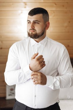 Beyaz gömlekli bir adam bir duvarın önünde duruyor. Kravat takıyor ve elleri cebinde.