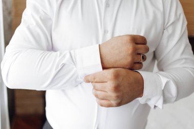 Beyaz gömlekli bir adam alyansını takmaya hazırlanıyor. O, gömleğini ve kollarını düzeltiyor ve elleri görünür durumda. Özel bir durum için hazırlık ve beklenti kavramı
