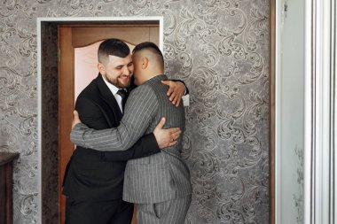 İki adam birbirine duvarları desenli bir odada sarılıyor. Adamlardan biri takım elbise ve kravat takmış.