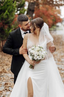 Bir gelin ve damat parkta fotoğraf için poz veriyorlar. Gelin bir buket çiçek tutuyor ve damat onu yanağından öpüyor. Sahne romantik ve mutlu.