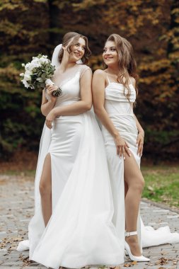 Beyaz gelinlik giymiş iki kadın fotoğraf için poz veriyor. İçlerinden biri elinde bir buket çiçek tutuyor.