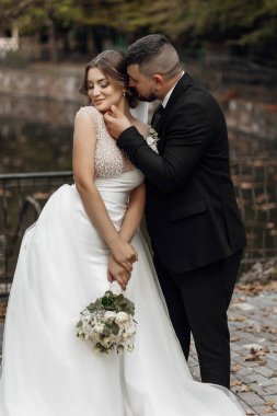 Bir gelin ve damat göletin önünde sarılıyorlar. Gelin elinde bir buket çiçek tutuyor.