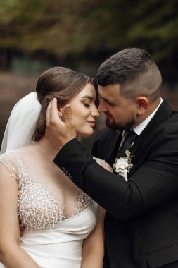 Bir gelin ve damat birbirlerinin yanaklarını öpüyor. Gelin beyaz bir peçe takıyor ve damat siyah bir takım giyiyor.