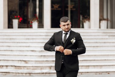 Takım elbiseli bir adam merdivenlerin üzerinde duruyor. O bir kravat ve siyah bir takım elbise giyiyor.