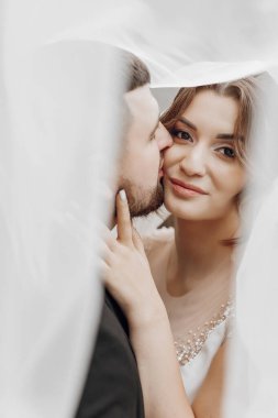 Bir gelin ve damat birbirlerinin dudaklarını öpüyor. Gelin beyaz bir elbise giyiyor ve damat siyah bir takım elbise giyiyor. Sahne samimi ve romantik, aşkı yakalıyor.