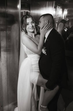 Asansörde bir gelin ve damat öpüşüyor. Gelin beyaz bir elbise giyiyor ve damat da takım elbise giyiyor.