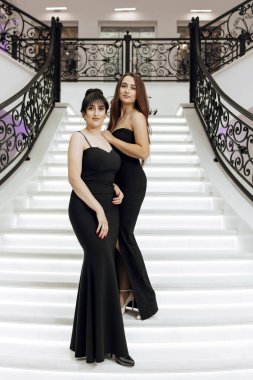 Siyah elbiseli iki kadın beyaz merdivende fotoğraf çektirmek için poz veriyor. Sahne zarif ve sofistike, çünkü kadınlar resmi giyinmiş ve merdiven güzel, süslü bir tasarım.