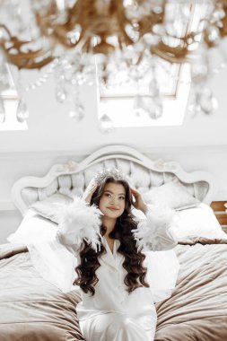 Taçlı ve beyaz cüppeli bir kadın bir yatakta oturuyor. Yatak kahverengi bir yorganla kaplı.