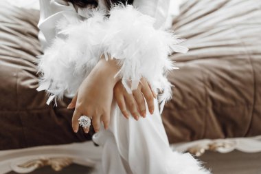 Bir kadın elleri kucağında bir yatağın üzerinde oturuyor. O, beyaz bir bornoz ve bir çift beyaz eldiven giyiyor. Eldivenler tüylerden yapılmış ve elmaslarla süslenmiş.