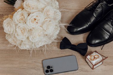 Masanın üzerinde bir buket beyaz gül bir cep telefonu ve bir çift siyah ayakkabının yanında duruyor.