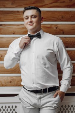 Beyaz gömlekli ve siyah papyonlu bir adam fotoğraf için poz veriyor. Tahta bir duvarın önünde duruyor.