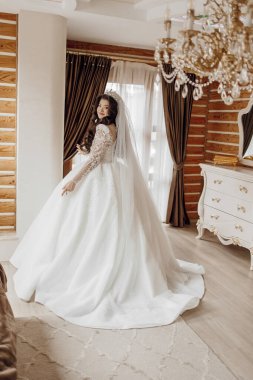 Beyaz elbiseli bir kadın pencerenin önünde duruyor. Bir duvak ve uzun beyaz bir elbise giyiyor.