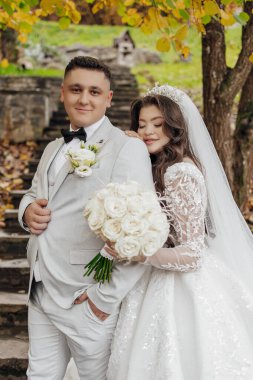 Bir gelin ve damat bir ağacın önünde fotoğraf için poz veriyorlar. Gelin beyaz bir elbise ve duvak giyerken damat takım elbise ve kravat takıyor. Bir buket beyaz gül tutuyorlar.