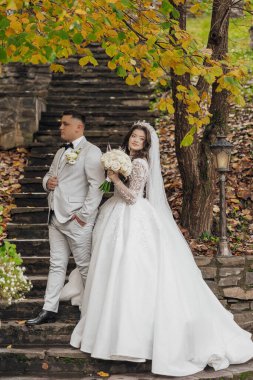 Bir gelin ve damat merdivende fotoğraf çektirmek için poz veriyorlar. Gelin beyaz bir elbise giyiyor ve damat bir takım elbise giyiyor. Gelin bir buket tutuyor ve damat da bir buket tutuyor.