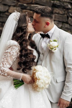 Taş duvarın önünde bir gelin ve damat öpüşüyor. Gelin beyaz bir elbise ve duvak giyerken damat takım elbise ve kravat takıyor. Gelin elinde bir buket çiçek tutuyor.