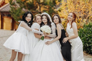 Bir grup kadın bir fotoğraf için poz veriyor, içlerinden biri gelinlik giyiyor. Gelin elinde bir buket çiçek tutuyor ve diğer kadınlar ona sarılıyor. Sahne mutlu ve kutlama dolu.