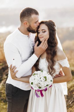 Bir gelin ve damat birbirlerine sarılıp öpüşüyorlar. Gelin elinde bir buket çiçek tutuyor.