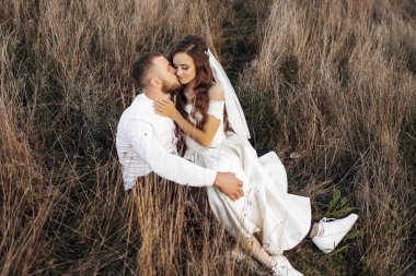 Bir gelin ve damat tarlada oturuyor, birbirlerini öpüyorlar. Sahne romantik ve samimi.