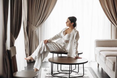 Beyaz elbiseli bir kadın oturma odasında bir masanın üzerinde oturuyor. Oda perdeler ve bir kanepeyle süslenmiş. Kadın, çevresinde rahat ve rahattır.