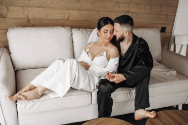 Bir adam ve bir kadın kanepede oturuyor, kadın beyaz bir elbise giyiyor. Adam kadının elini tutuyor.