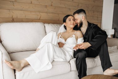 Bir adam ve bir kadın kanepede oturuyor, kadın beyaz bir elbise giyiyor. Adam onun elini tutuyor.