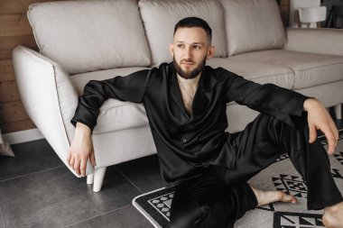 Siyah takım elbiseli ve siyah pantolonlu bir adam oturma odasında bir halının üzerinde oturuyor. Gülümsüyor ve rahatladı.