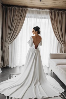 Beyaz elbiseli bir kadın pencerenin önünde duruyor ve dünyaya bakıyor. Elbise uzun ve dalgalı, ve kadının saçı topuz şeklinde geri çekiliyor. Sahne huzurlu ve huzurlu.