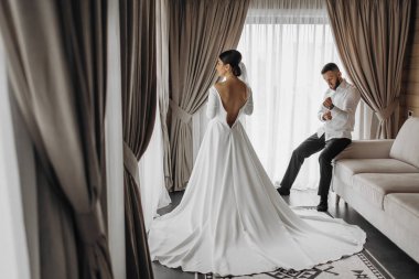 Bir gelin ve damat düğünleri için hazırlanıyorlar. Gelin beyaz bir elbise giyiyor ve damat siyah bir takım elbise giyiyor. Kanepenin ve perdenin önünde duruyorlar.