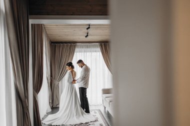 Bir gelin ve damat düğünleri için hazırlanıyorlar. Gelin beyaz bir elbise giyiyor ve damat beyaz bir gömlek giyiyor. Kanepenin ve pencerenin önünde duruyorlar.