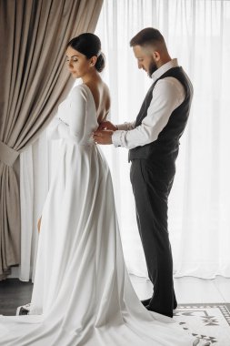 Gelin ve damat bir perdenin önünde, gelinle birlikte uzun beyaz bir elbise giyiyorlar.