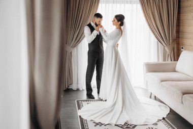 Gelin ve damat bir kanepenin önünde duruyorlar, gelin beyaz bir elbise giyiyor.