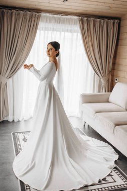 Beyaz elbiseli bir kadın kanepenin önünde duruyor. Kanepe beyaz ve üzerinde bir desen var. Oda perdeler ve halıyla süslenmiş.