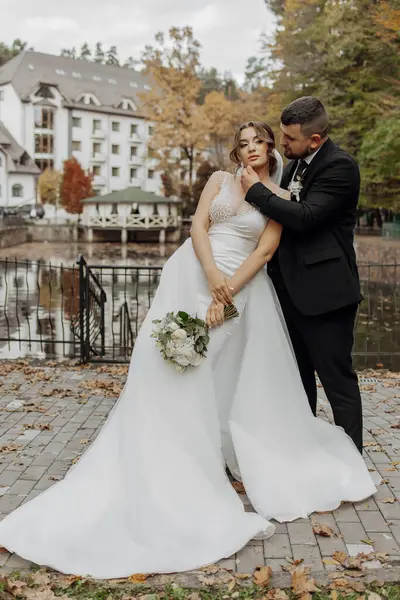 Bir gelin ve damat binanın önünde fotoğraf için poz veriyorlar. Gelin beyaz bir elbise giyiyor ve damat takım elbise giyerken elinde bir buket tutuyor. Sahne romantik ve mutlu.