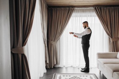 Takım elbiseli bir adam pencerenin önünde duruyor, kravatını düzeltiyor. Oda perdeler ve halı ile süslenmiş, rahat ve rahat bir atmosfer sağlıyor.